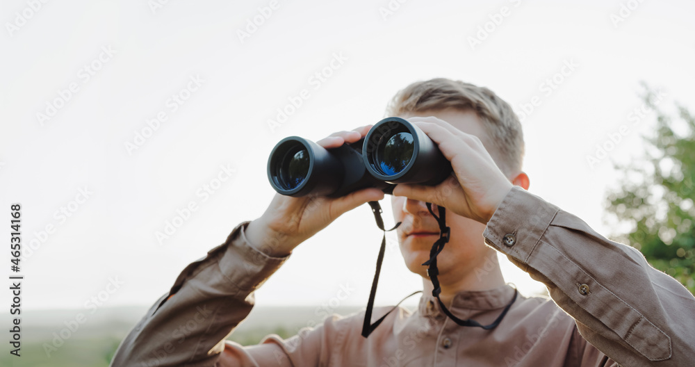 Fototapeta premium A young man looks through binoculars at a beautiful hilly landscape.The concept of hunting, travel and outdoor recreation. Banner with copy space.A traveler or hunter is observing through binoculars.