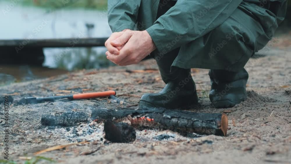 A man in a raincoat warms his hands over smoldering firewood on an autumn day on the sandy shore of the reservoir. Hands and a bonfire close-up. A man in a raincoat warms his hands over smoldering firewood on an autumn day on the sandy shore of the reservoir. Hands and a bonfire close-up.