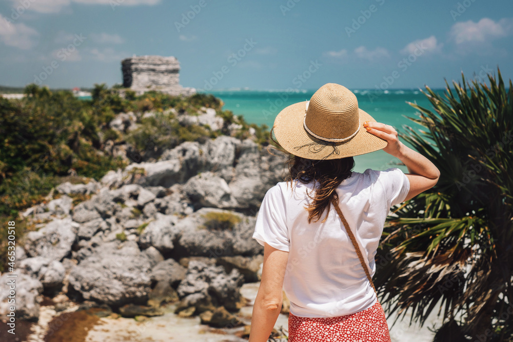 A romantic girl tourist in a hat stands on a cliff overlooking the Tulum ruins and the Caribbean Sea. Young woman view from the back against the backdrop of the ruins of Tulum, Mexico. Popular place