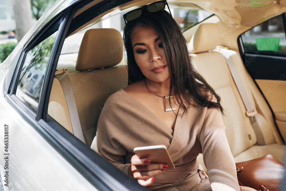 Serious beautiful young Asian woman with sunglasses on head sitting into expensive car and checking messengers on phone Serious beautiful young Asian woman with sunglasses on head sitting into expensive car and checking messengers on phone