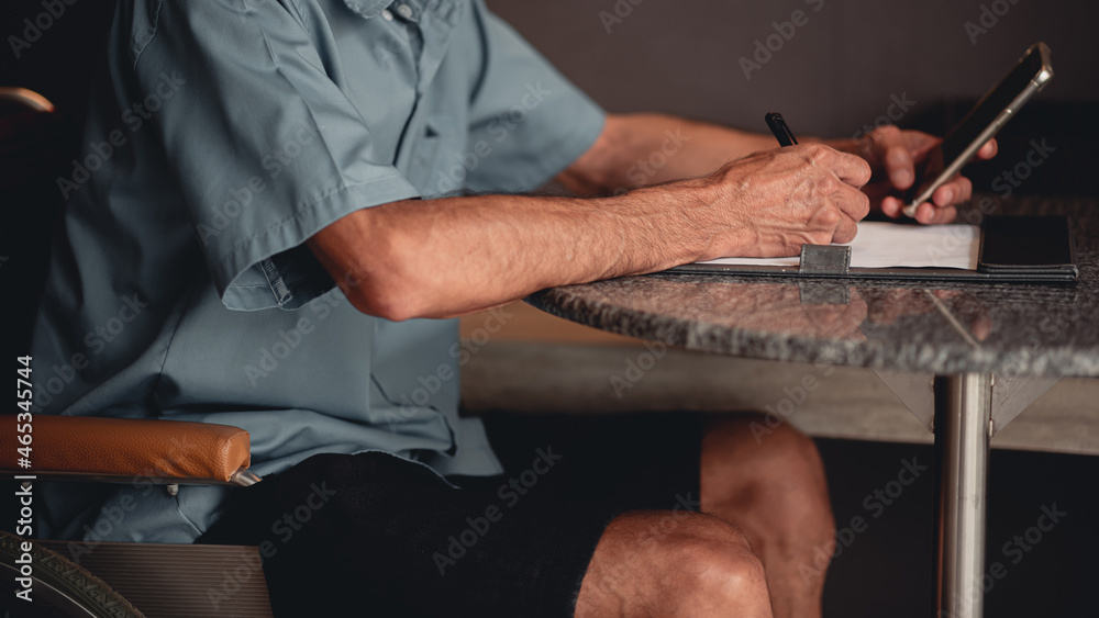 Fototapeta premium Hands of senior man with disability holding a pen to writing something with business working or sign name on important document by himself on desk in home office, People work and using phone concept.