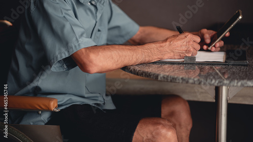 Hands of senior man with disability holding a pen to writing something with business working or sign name on important document by himself on desk in home office  People work and using phone concept.