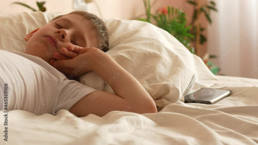 A Caucasian boy of 7-9 years old wakes up in the daylight from a smartphone alarm in his bed at home, turns off the signal and falls asleep again. the child does not want to wake up in the morning A Caucasian boy of 7-9 years old wakes up in the daylight from a smartphone alarm in his bed at home, turns off the signal and falls asleep again. the child does not want to wake up in the morning