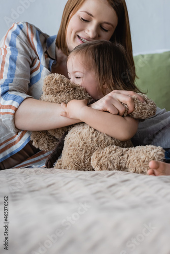 Smiling woman hugging daughter with down syndrome and soft toy on bed.