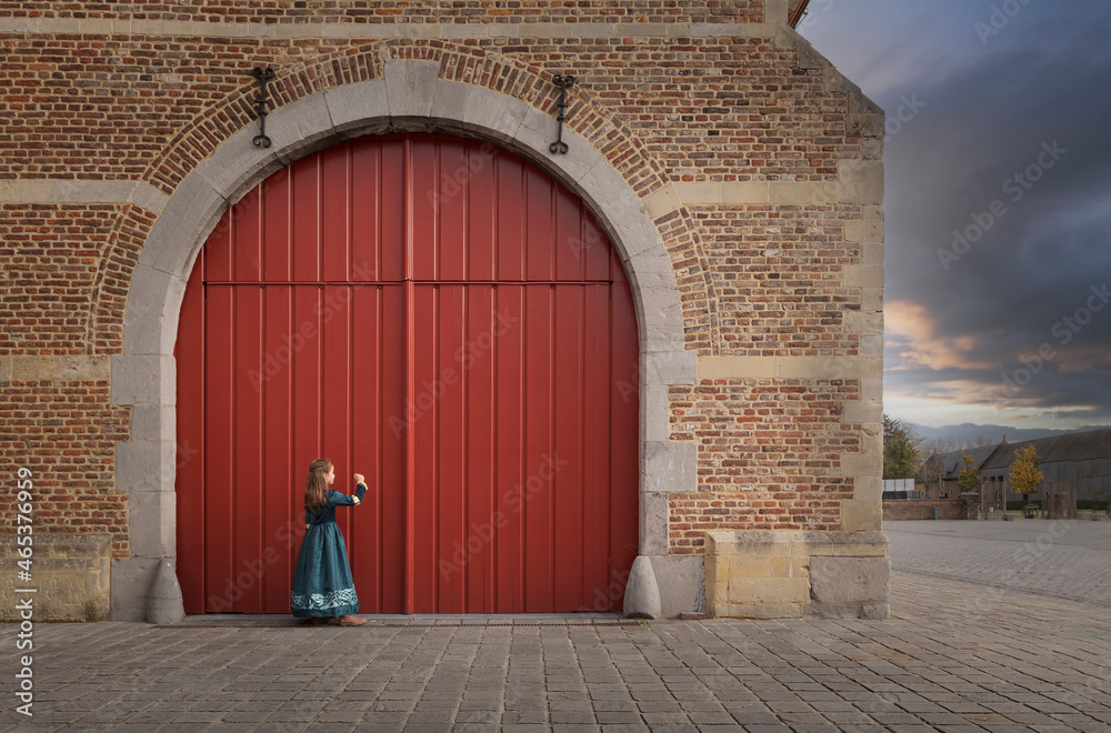 Little girl in medieval dress knocking on the big red gate of the old castle