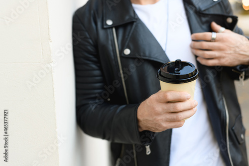 Cropped view of coffee to go in hand of blurred man in leather jacket near white wall 