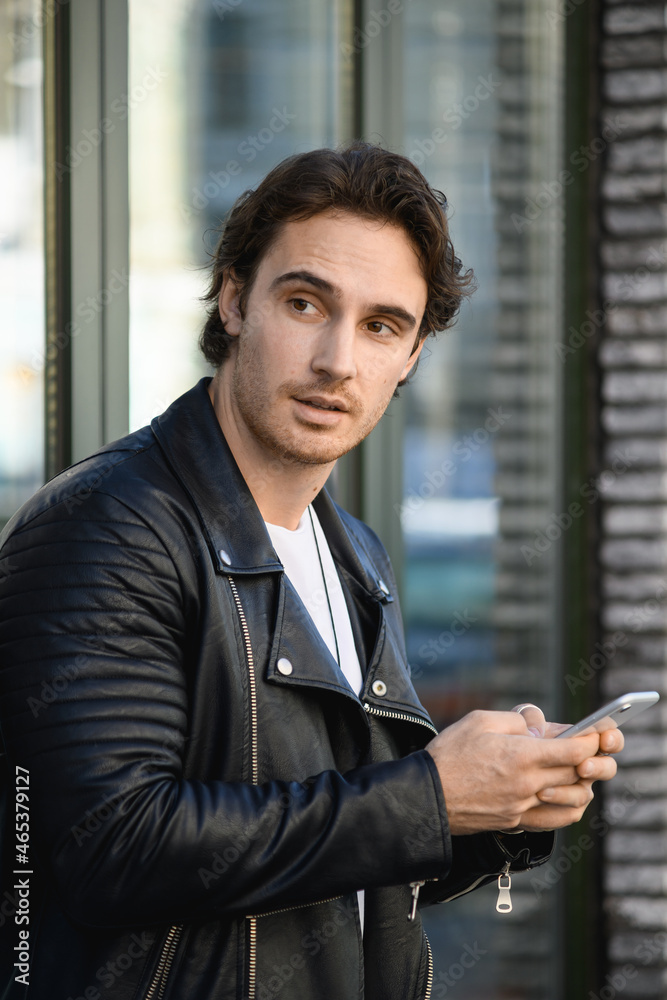 Fototapeta premium Fashionable man in leather jacket using smartphone on city street 