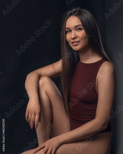 Studio photo of pretty brunette woman sitting on cube and hugging her leg with hand on black background © Elena Odareeva