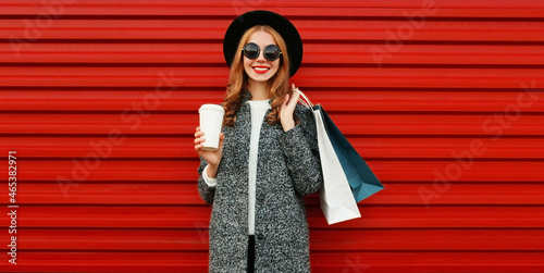 Portrait of beautiful smiling woman with shopping bags wearing gray coat, hat on colorful red background © rohappy