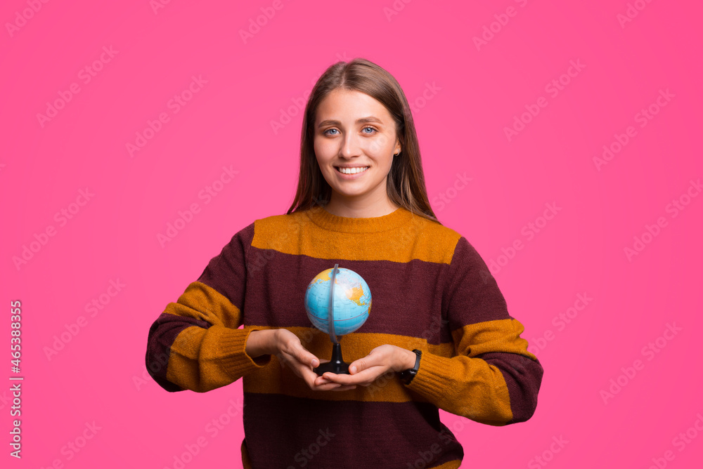 Smiling female student is holding a little Earth globe while standing over pink background. Smiling female student is holding a little Earth globe while standing over pink background.