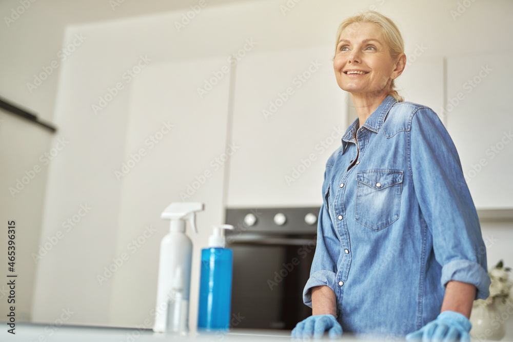 Fototapeta premium Attractive mature woman in rubber gloves smiling away, ready for clean up in the kitchen. Detergents, sanitizer spray and gel in bottle on the surface