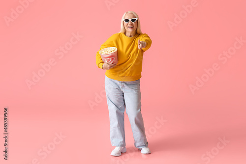 Mature woman with tasty popcorn pointing at viewer on color background © Pixel-Shot