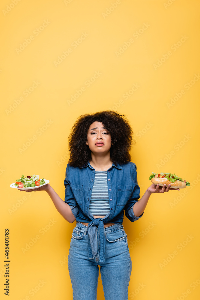 sad african american woman with vegetable salad and hot dog looking at camera isolated on yellow sad african american woman with vegetable salad and hot dog looking at camera isolated on yellow