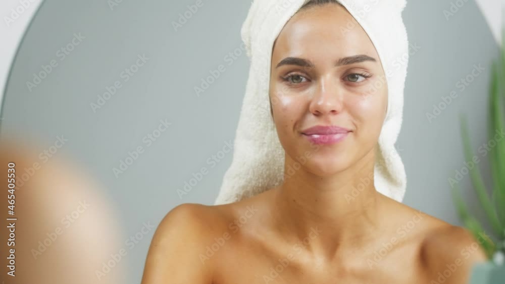 Woman using moisturizing lipstick and smiling, morning skincare routine. Young female student with bare shoulders wearing towel on head looking in mirror in spa salon. Natural day make up concept.
