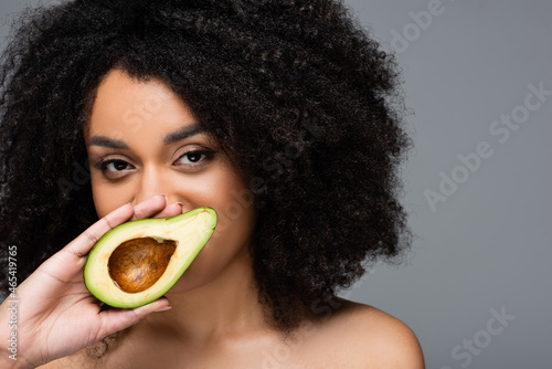 pretty african american woman holding half of fresh avocado near face isolated on grey