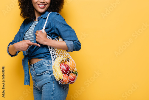 cropped view of smiling african american woman pointing at string bag with ripe fruits isolated on yellow photo