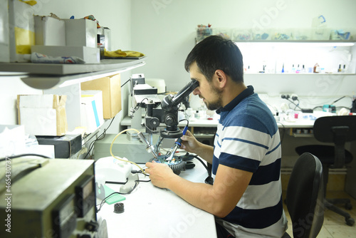 A male repairman in the workshop looks at the microcircuit of an electrical device through a microscope while doing electrical soldering of equipment