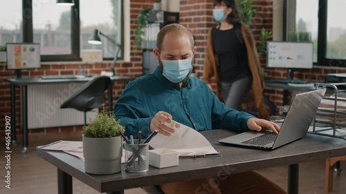 Entrepreneur looking at screen and working with laptop to design rate charts on papers for project planning at desk. Man using computer to plan marketing strategy and wearing face mask. photo