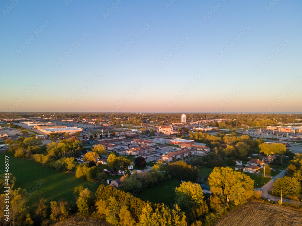 aerial view of urban city in autumn with bright blue sky and colored foliage.  water tower seen in distance.  neighborhood marked by rows of trees. blue sky in horizon with pink tinges of sun.      
