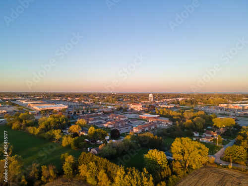 aerial view of urban city in autumn with bright blue sky and colored foliage. water tower seen in distance. neighborhood marked by rows of trees. blue sky in horizon with pink tinges of sun. 