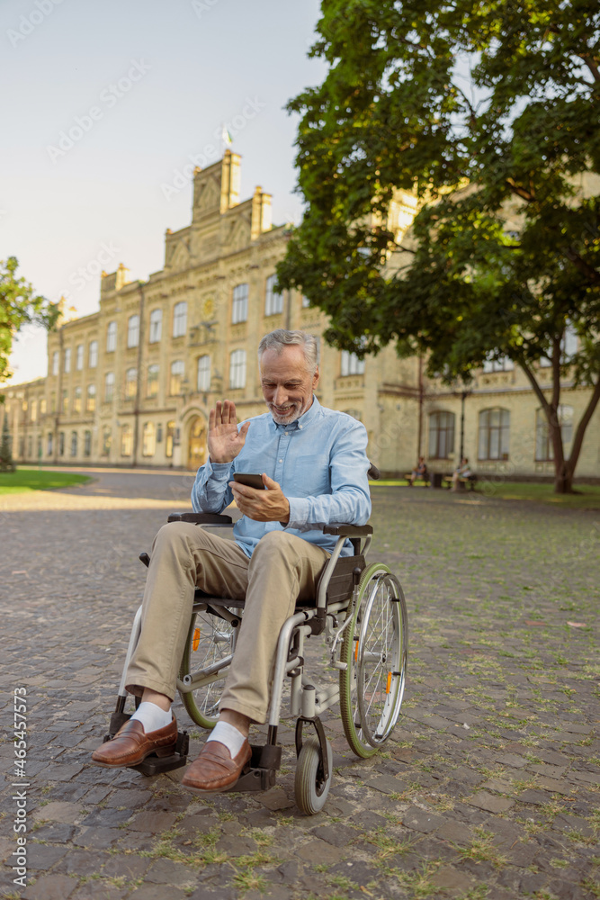 Smiling senior man, recovering male patient in wheelchair making a video call using smartphone while resting alone in the park near hospital