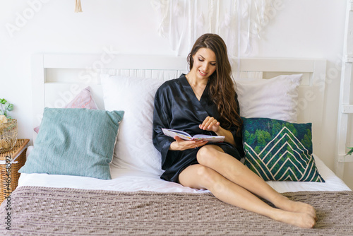 Beautiful girl lies on white bedding at home. A young woman in a dark gray silk robe is reading a book. Caucasian brunette lady is enjoying the morning in a comfortable bed. Soft focus.