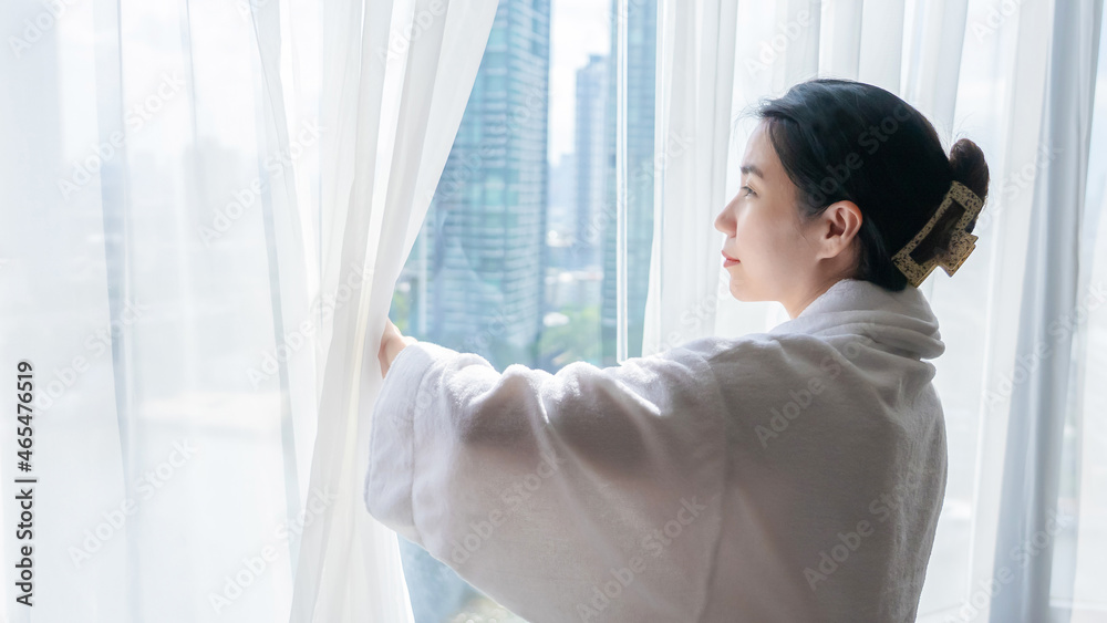 Confident young woman wear nightgown stand open curtain lace looking outside enjoy morning view. Lady wearing shower robe opens white window curtains and looks at the skyscrapers in the big city. Confident young woman wear nightgown stand open curtain lace looking outside enjoy morning view. Lady wearing shower robe opens white window curtains and looks at the skyscrapers in the big city.