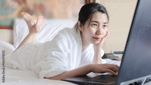 asian woman relaxing in the hotel and working with laptop on a bed after a shower with bathrobe. Freeland asia girl work on bed from hotel comfortably smiling enjoying care free time holiday #465476535