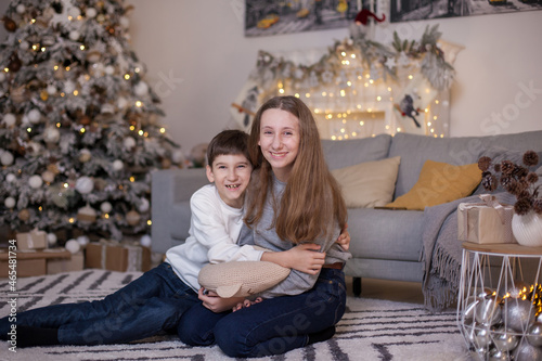 Happy brother and sister sitting on floor and fir tree with presents and looking at camera. Boy and girl at Christmas time. Merry Christmas and Happy New Year. Family values. Waiting for a holiday. © pijav4uk
