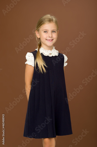 Adorable blonde wearing in white blouse and blue dress standing against brown background.