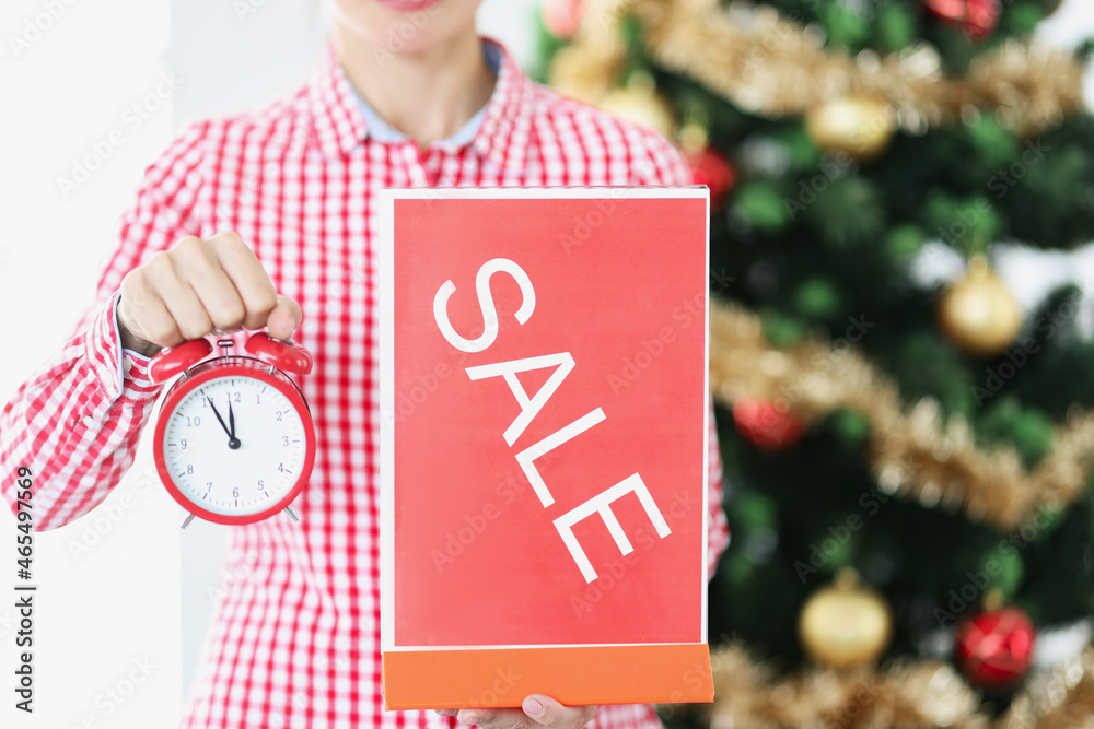 Woman holding red alarm clock and sale plate near new year tree closeup Woman holding red alarm clock and sale plate near new year tree closeup
