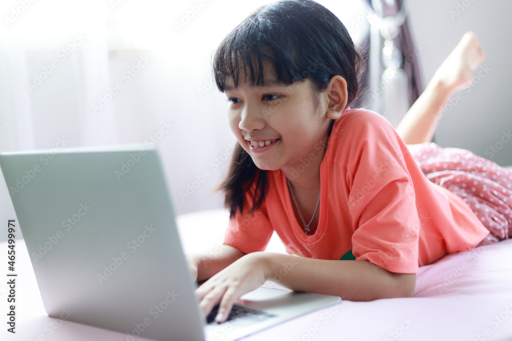 Portrait shot little Asian girl using a laptop computer to learn from home and laying on the bed with happiness, self-quarantine and self-education concept, selective focus shallow depth of field Portrait shot little Asian girl using a laptop computer to learn from home and laying on the bed with happiness, self-quarantine and self-education concept, selective focus shallow depth of field