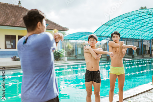 two teenage swimmers in swimming trunks doing arm stretching with trainer