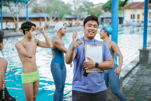 a swimming trainer smiles with an ok finger gesture while holding a clipboard