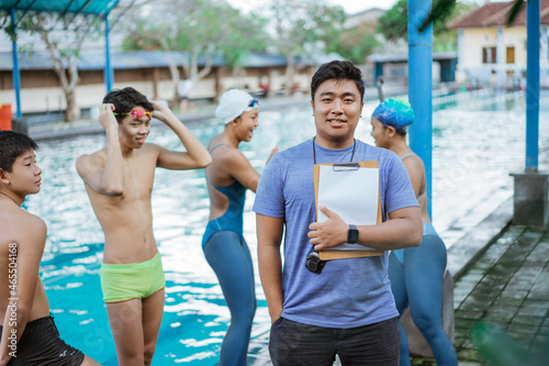 a swimming trainer smiling with one hand in pocket while holding a clipboard with his students