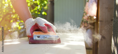 A man polishes a wooden surface with sandpaper. A male hand in protective gloves works outdoors. Sawdust dust glows in the sun. Horizontal format, close-up, side view Layout with space for text.