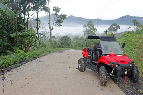 A red utility terrain vehicle (UTV) parked at the side of a country road with tropical trees and mist covered mountains in the background