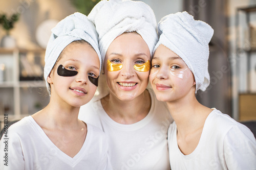 Portrait of caucasian mother and her two daughter posing at home with collagen patches under eyes and bath towel on head. Concept of family  skin care and domestic lifestyles.