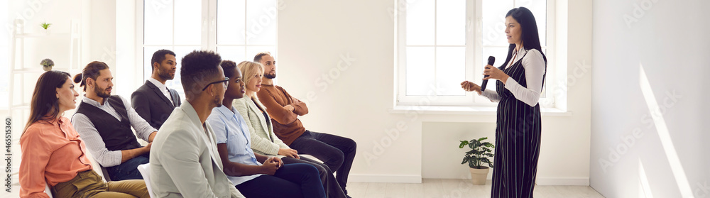 Experienced talented speaker making speech for multiracial audience. Woman standing with microphone and sharing knowledge and skills with multiethnic group of people, side view. Website banner, header Experienced talented speaker making speech for multiracial audience. Woman standing with microphone and sharing knowledge and skills with multiethnic group of people, side view. Website banner, header