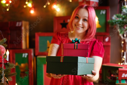 Selective focus. A woman with pink hair holds out a red gift in a beautifully decorated room for Christmas