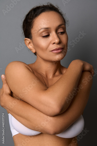 Close-up beauty portrait of self-confident middle aged African woman with perfect clean shiny healthy skin, hugging herself, isolated over gray background with copy space. Body and skin care concept