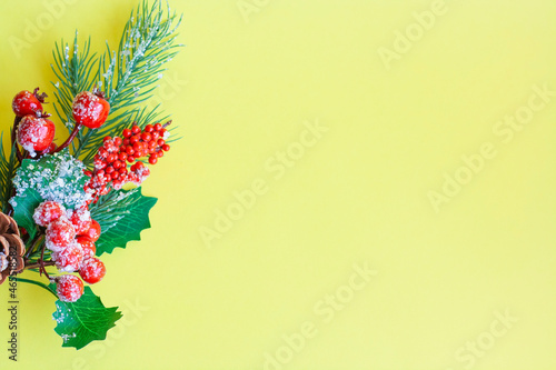 Christmas tree twig with red berries and holly leaves on a yellow background, copy space