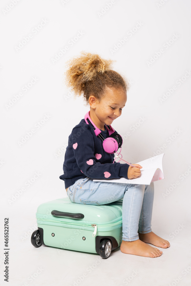 Beautiful little girl sitting on a suitcase and holding a notebook isolated over white background. Beautiful little girl sitting on a suitcase and holding a notebook isolated over white background.
