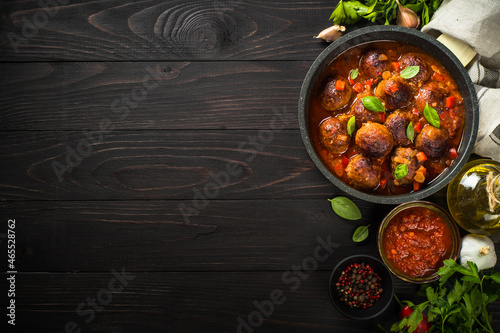 Meatballs in tomato sauce in a skillet on dark wooden table with ingredients. Top view with copy space.