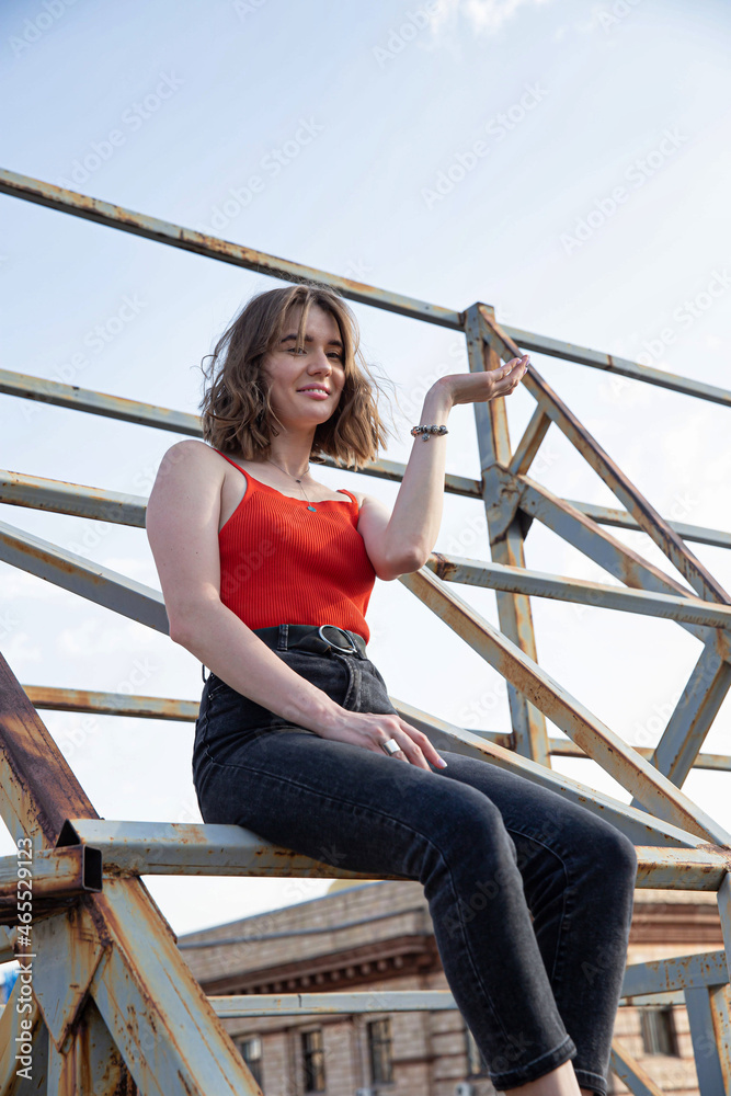 Smiling young european woman in red top and dark jeans sitting on metal construction