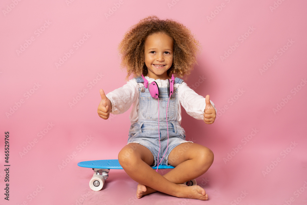 Cool little toddler girl sits on a skateboard and looking at the camera with thumbs up isolated over pink background Cool little toddler girl sits on a skateboard and looking at the camera with thumbs up isolated over pink background