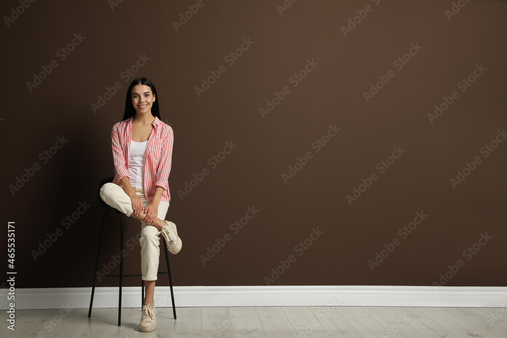Beautiful young woman sitting on stool near brown wall. Space for text Beautiful young woman sitting on stool near brown wall. Space for text