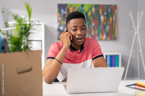 A customer service employee is sitting in an office at desk in front of laptop screen, talking on phone, trying to solve caller's problem checking available dates taking complaints, his face disgusted