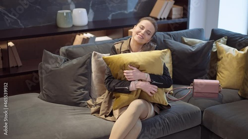 Portrait of smiling happy Caucasian slim beautiful woman admiring and hugging decorative mustard cushion sitting on couch in furniture showroom. Attractive female buyer making choice in shop photo