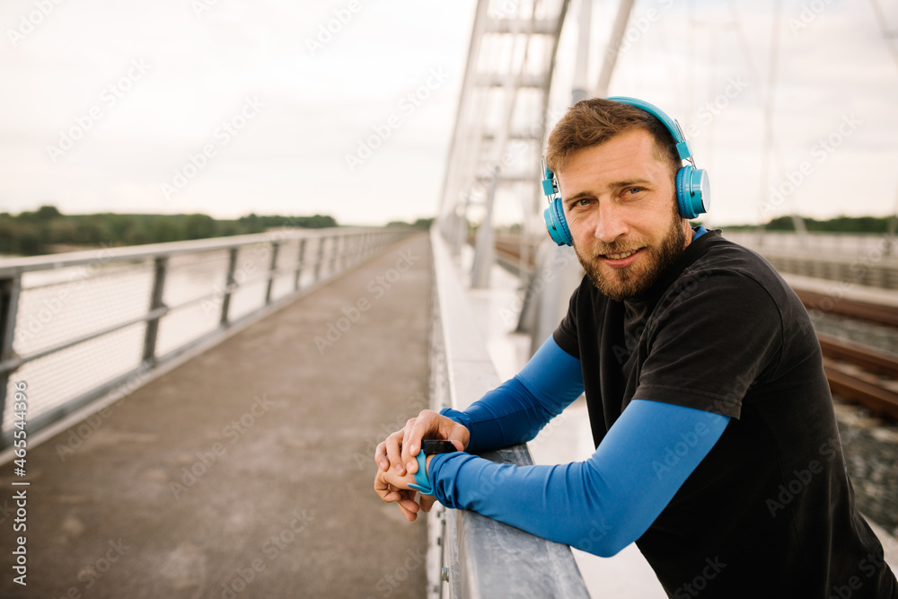 Runner man standing on a bridge taking a break after running early in the morning listening to music on his blue headphones
